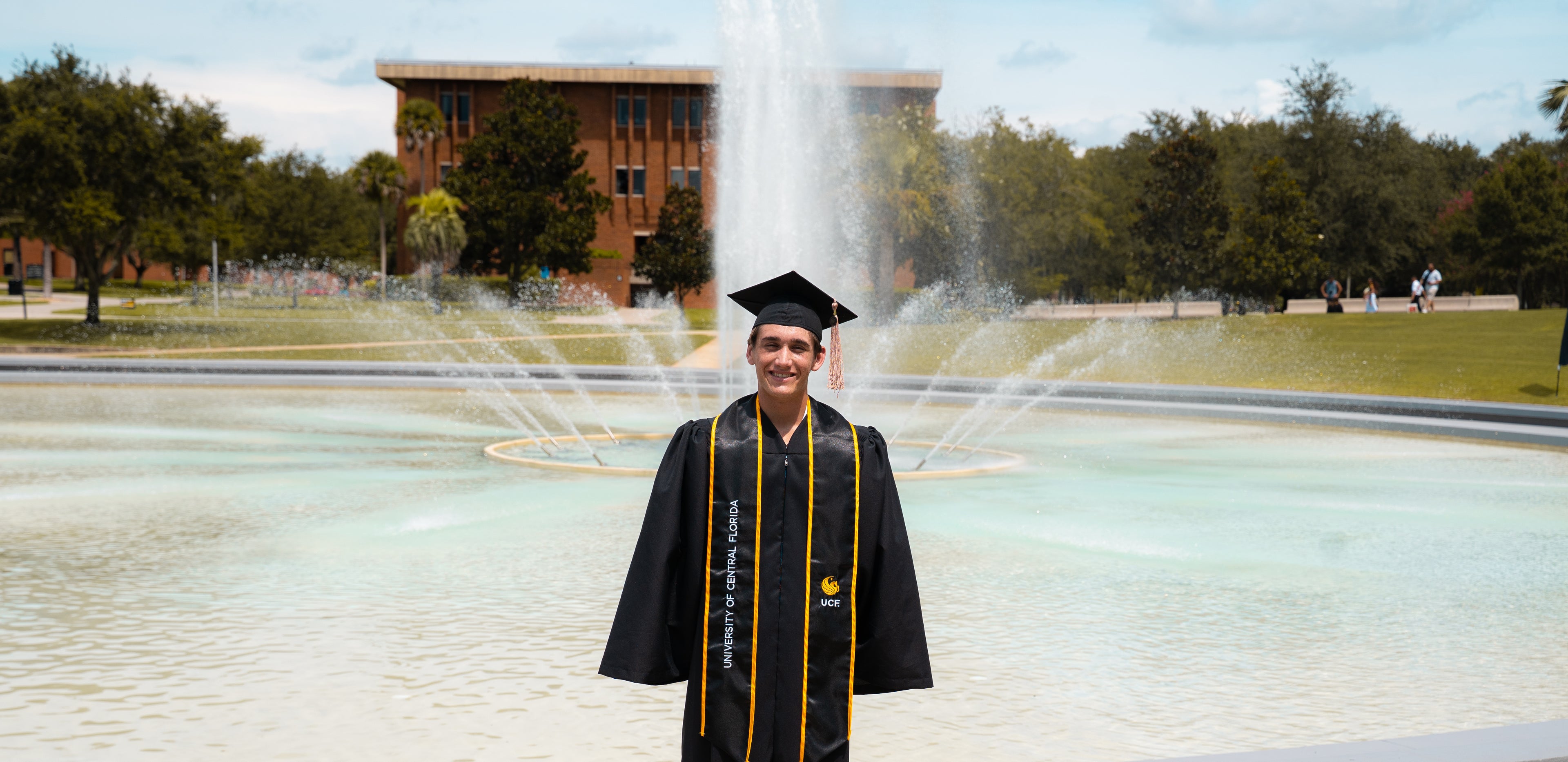 Graduate in cap and gown standing in front of a fountain on a college campus